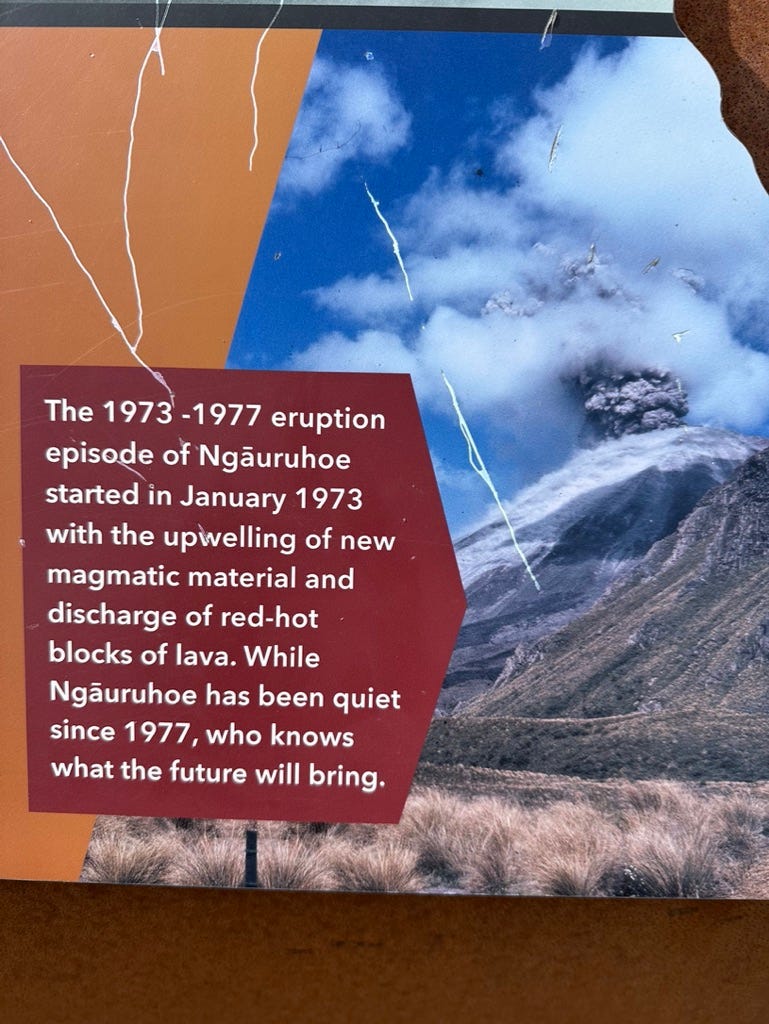 Sign with photo of volcano. Reads: “The 1973–1977 eruption episode of Ngauruhoe started in January 1973 with the upwelling of new magmatic material and discharge of red-hot blocks of lava. While Ngâuruhoe has been quiet since 1977, who knows what the future will bring.”