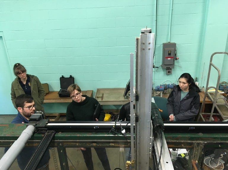 Students stand with staff members looking at an upright the metal mechanism that makes up a fish passage model.