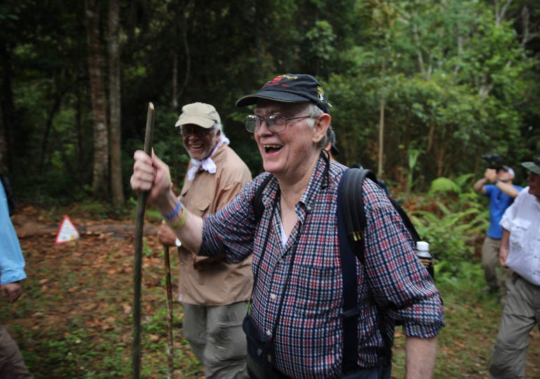 Two smiling men walking through the jungle with walking sticks.