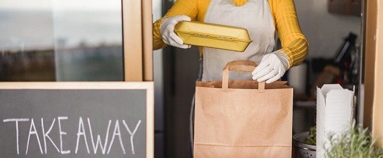 A photograph of a store window with a chalkboard that reads ‘takeaway’. Through the window is a person, who can be seen from the neck to the hip, putting a yellow take away container into a brown paper carry bag.