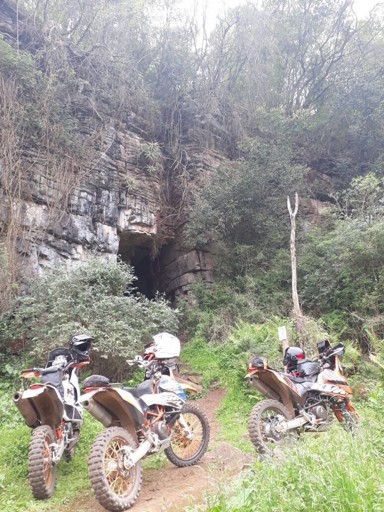 Three scrambler motorcycles in front of a cave inside a rock cliff in a valley
