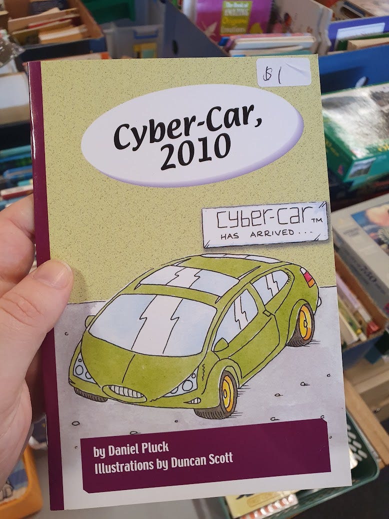 Photo of a hand Holding the book titled “Cyber Car 2010”. The cover has an illustration of a green sedan with the sign “cyber car has arrived” in the background. The book is by Daniel Pluck. Ilustrations by Duncan Scott