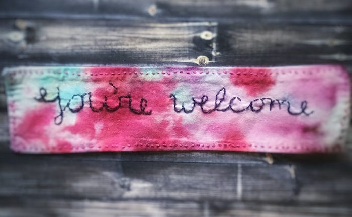 Photograph of a tie dyed piece of fabric with the words “You’re welcome” stitched on it. It was ironed onto a cloth mask.