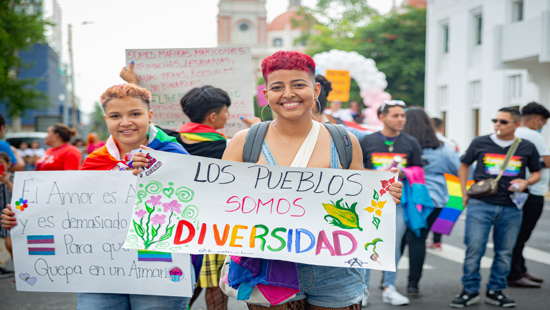 People participate in an LGBTQI+ walk in Honduras. A marcher at the front of the group smiles while holding a sign that reads “Los Pueblos Somos Diversidad.”