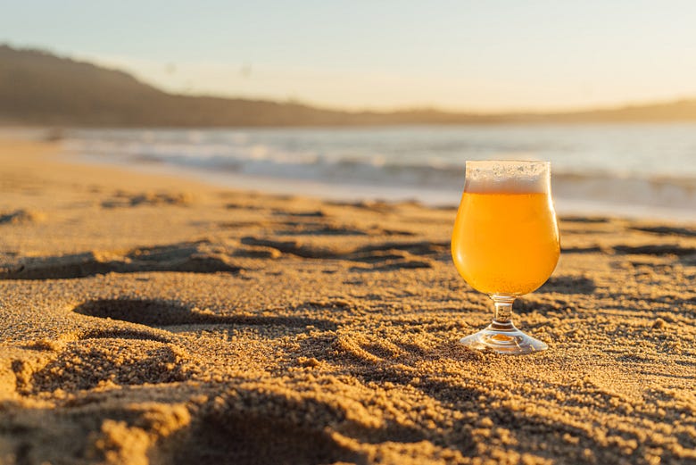 A tall glass of beer sitting on a beach in front of the ocean.