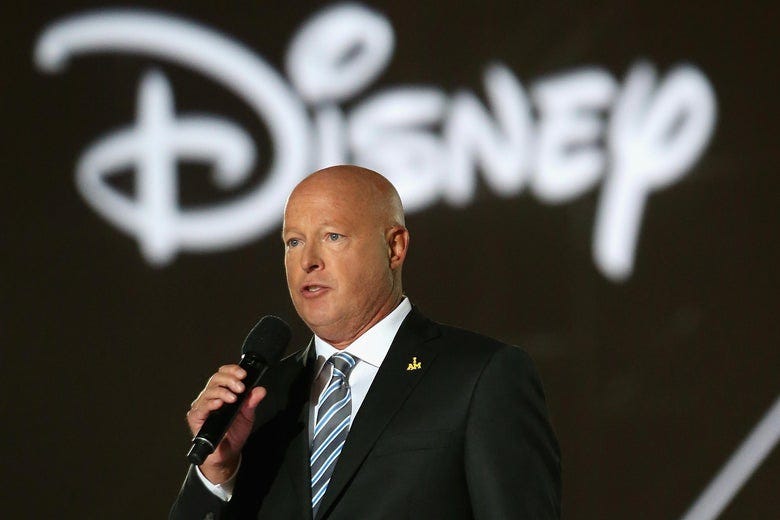 A photo of Disney CEO — Bob Chapek — wearing  a black suit with a white dress shirt and blue-and-black striped tie, holding a microphone, standing in front of a Disney logo.