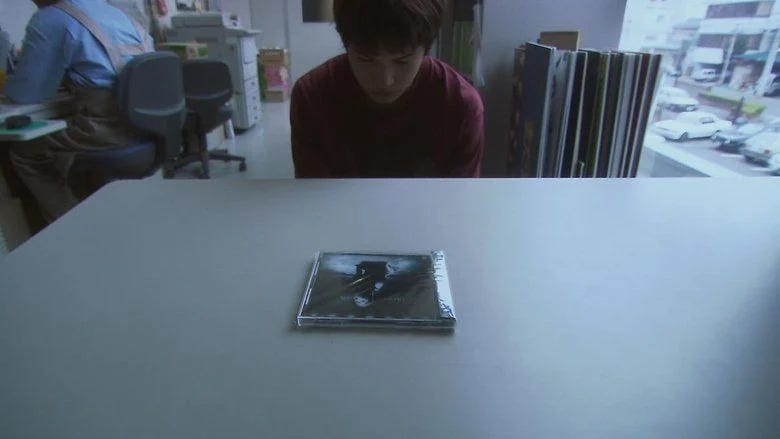 A young teen boy with an embarrassed/defeated expression and posture staring down at a CD lying on a desk in front of him.