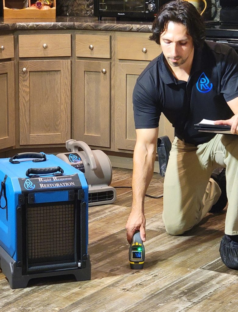 Restoration specialist from Rapid Response Restoration assessing crawl space moisture levels using a digital hygrometer on wooden flooring, with professional dehumidifier equipment in the background, ensuring optimal crawl space health and maintenance.