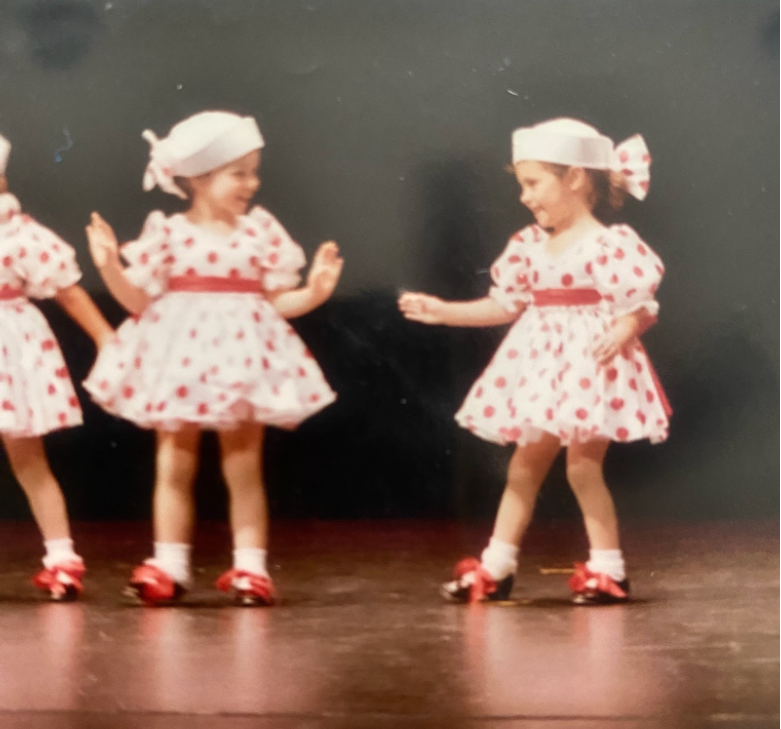 A young Riley Young stands on stage wearing a white dress with red polka dots, two other fellow dancers stand beside her