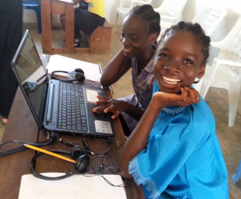 Two girls share a computer to learn in Kolibri. Sitting at a desk, one stares at the screen while the other smiles at the camera. There are notebooks, pencils and headphones on the table.