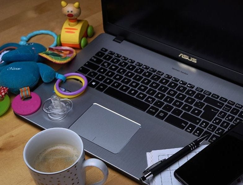 An openlaptop surrounded by children’s toys, with a cup of coffee in front of it and a pen, diary and phone resting on the left hand side