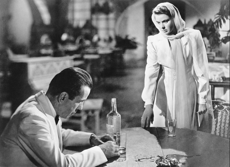 A man in a tuxedo sulks while drinking and listening to a woman dressed in white.