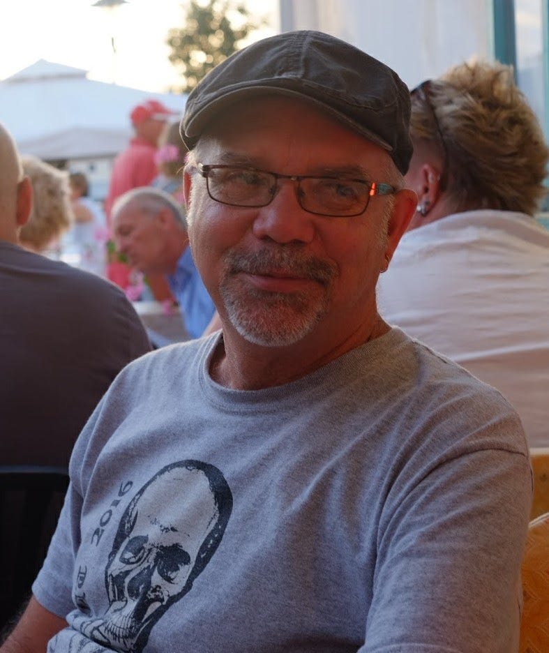A man in a hat is seated outside with others. He has on glasses and a t-shirt and is looking to the right.