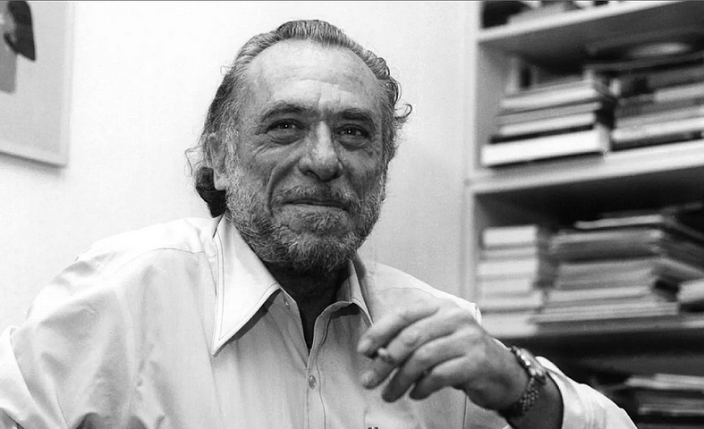 Charles Bukowski smoking a cigarette in front of a pile of books