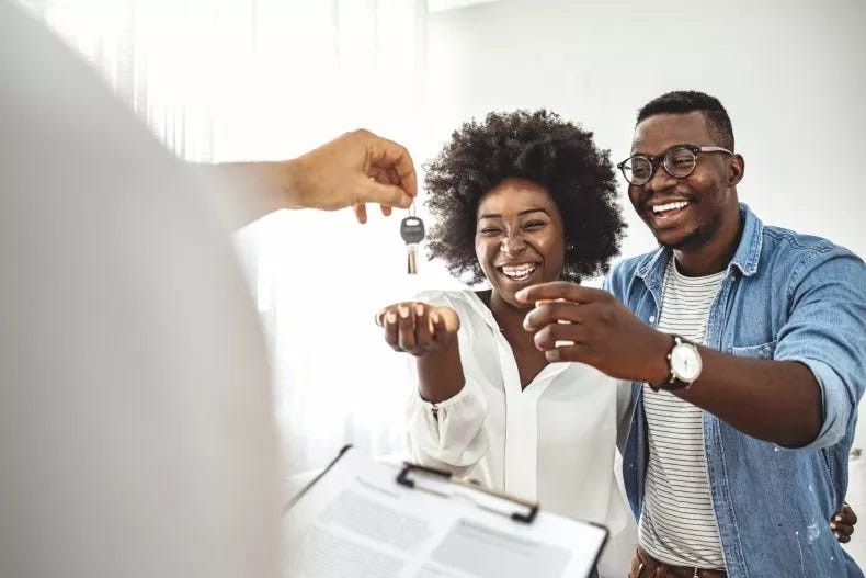 Casal, mulher negra de cabelos escuros e curtos usando uma camisa branca e sorrindo estendendo a mão para pegar uma chave na mão de outra pessoa. Homem negro usando óculos camisa jeans aberta e uma camiseta listrada, cabelos escuros e curtos e sorrindo. Ambos em frente a um contrato.