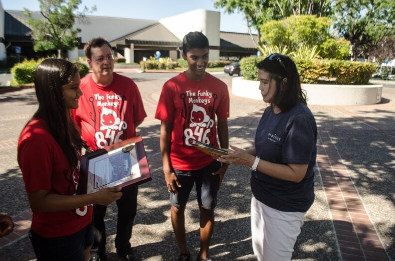 David Giandomenico, Shikhar Jagadeesh, and Ria Pradeep present a team photo and photojournal to an employee of Intuitive Surgical.