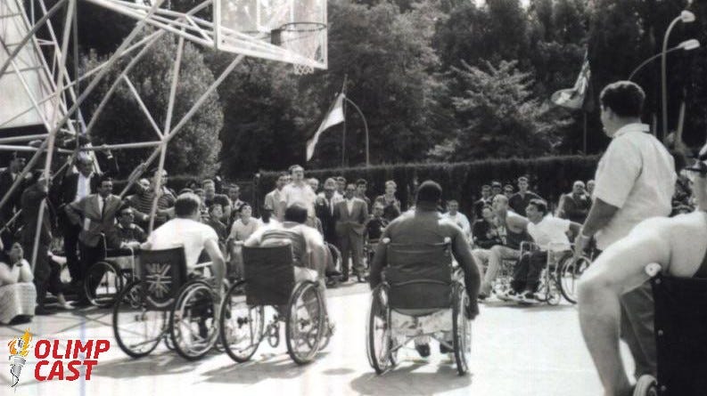 Três homens jogam basquete em cadeira de rodas durante os Jogos Paralímpicos de Roma-1960. A imagem é em preto e branco. Os jogadores estão de costas, numa quadra que tem uma tabela e um aro de basquete adaptados para uma altura mais baixa, aparecendo no alto da imagem. Ao redor da quadra há um número grande de pessoas em pé observando a partida, que parece acontecer num parque — há árvores no fundo da imagem.
