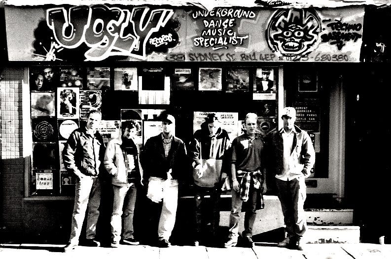 Six men in casual clothes standing outside record shop in Brighton called ‘Ugly’ — the shop is decorated in grafitti and the photo is in black and white