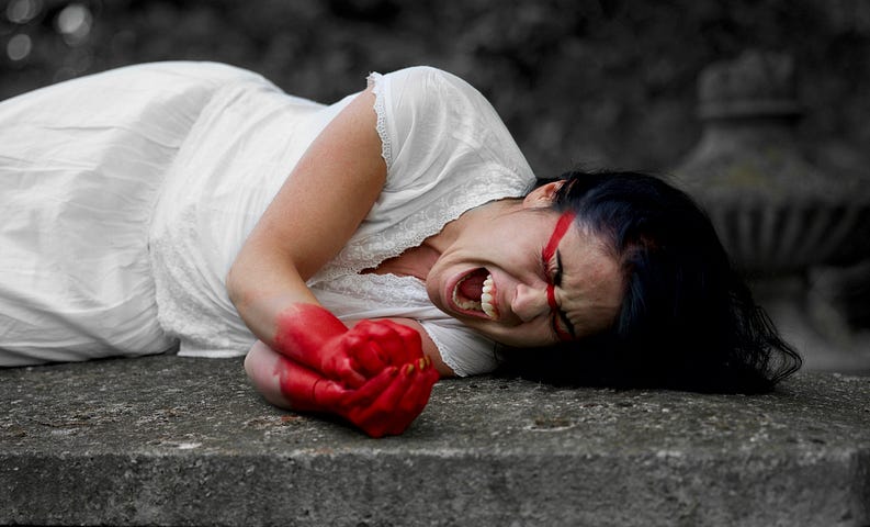 A woman with black hair wearing white and laying on a rock with red hands and face