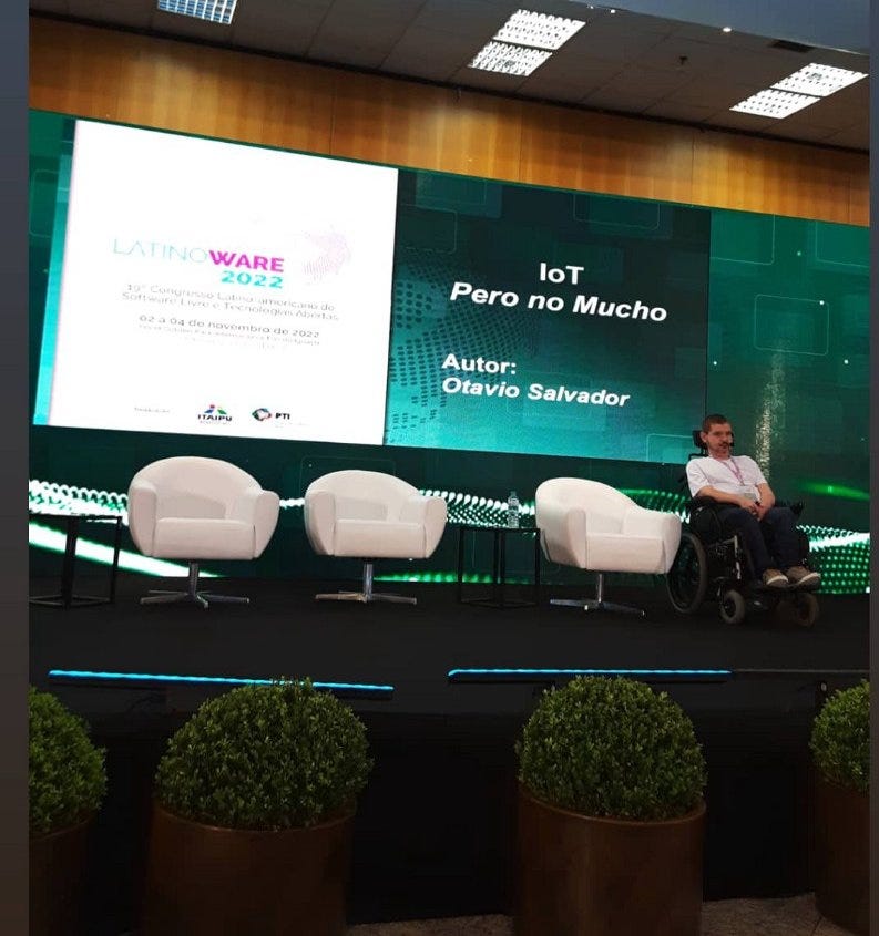 Otavio Salvador aparece no palco de camiseta branca com a logomarca do ShellHub e calça jeans preta em frente a uma tela com a extensão de todo o palco onde é reproduzido o slide de capa de sua palestra com o título: IoT pero no mucho.