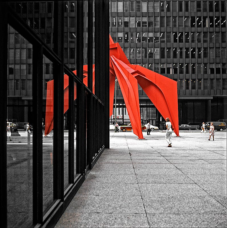 Half of a large red metal sculpture appears around the corner of a modern skyscraper, creating an illusion of symmetry, with that part of the red metal sculpture reflected in the building’s windows. A handful of relatively small people walk around in the middle distance.