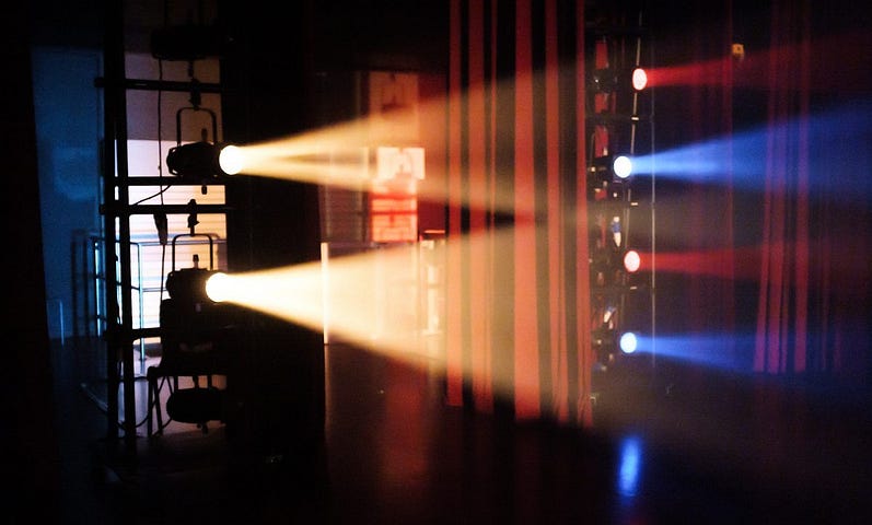 Backstage theater lights with a theater curtain in the background. To the left, two big yellow lights project horizontally towards the right. To the right, smaller alternated red and blue lights also project towards the right.