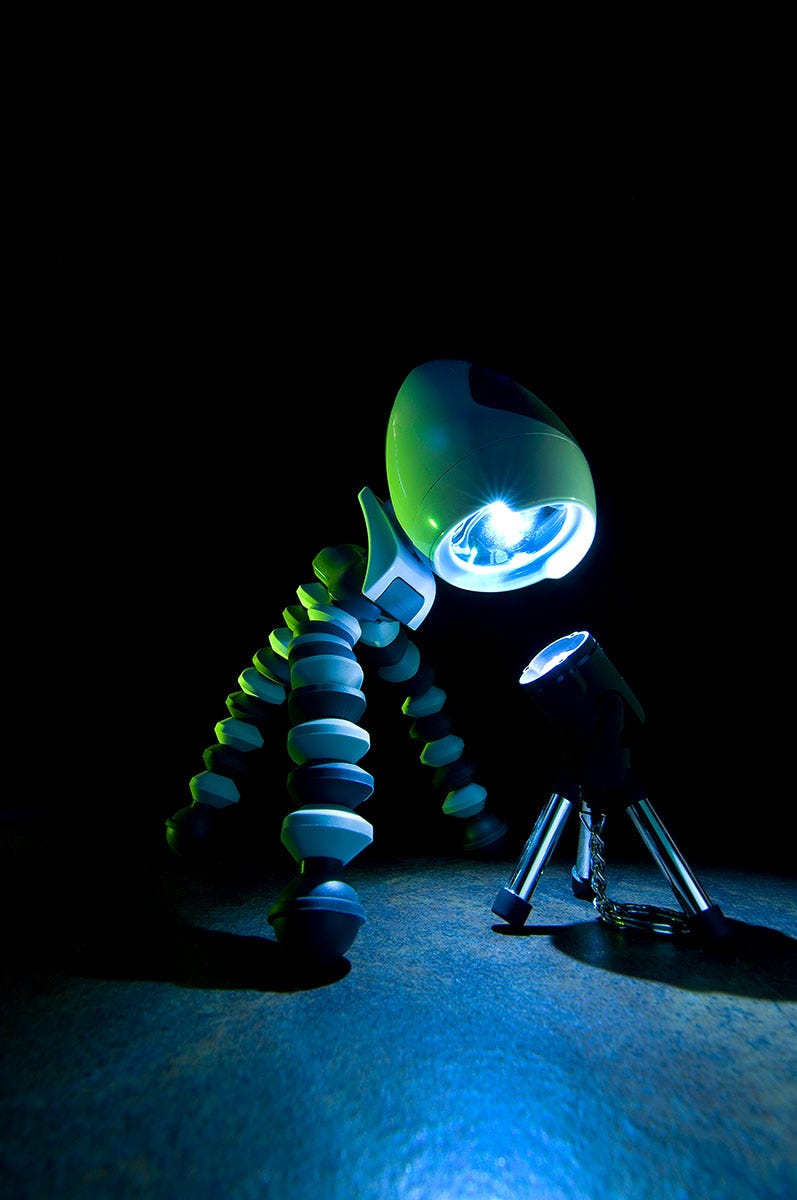 A large 3-legged flashlight looms over and shines its brighter light down onto a smaller 3-legged light, in an attempt to dominate and intimidate its rival.