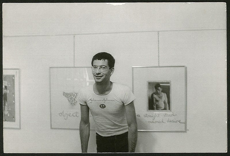 A black and white photo of David McDiarmid at his first solo exhibition. A print of Marlon Brando behind him is captioned ‘a straight stud named desire’.