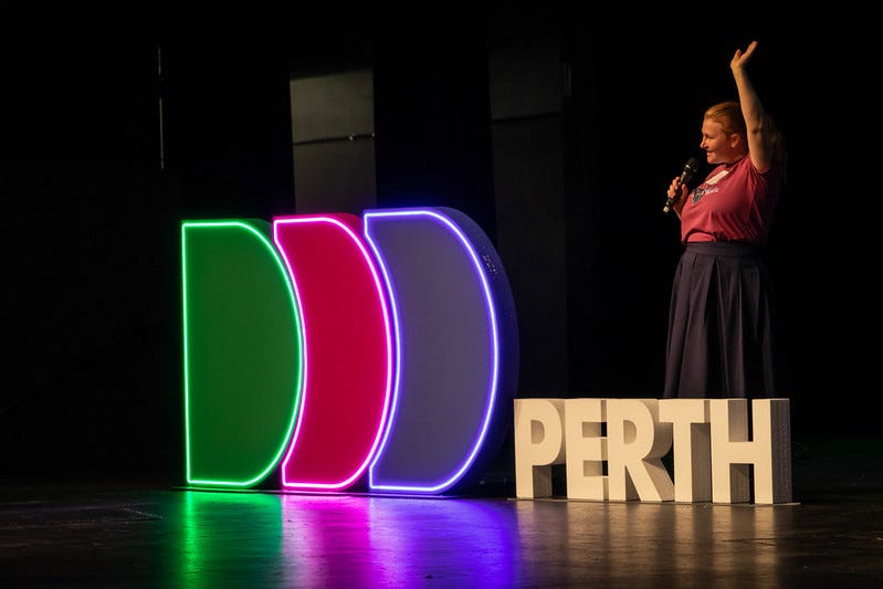 Rebecca standing behind the neon DDD Perth sign on the main stage of DDD Perth 2022. She has one hand in the air and is holding a microphone to her mouth with her other hand.