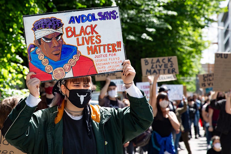 A white woman at a march holds up a sign with a Black comic character that reads, “Avdol Says: Black Lives Matter!”