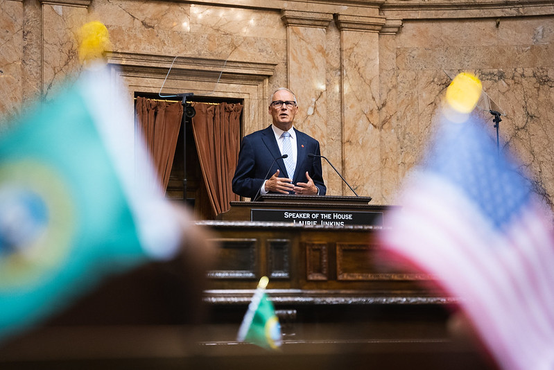 Photo of Jay Inslee in the House chambers speaking from the rostrum. Blurred images of the Washington state and U.S. flags frame the foreground.