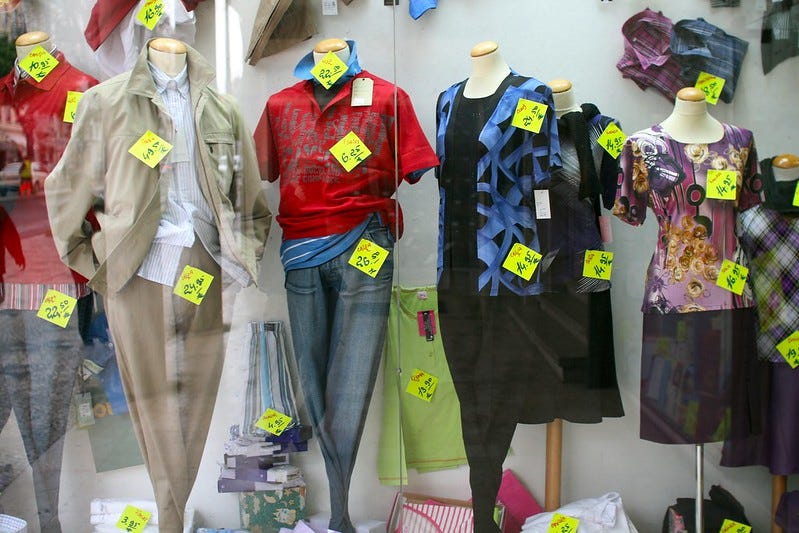 A department store window display of several headless mannequins wearing clothes that are covered in neon yellow price tags that look like post-it notes