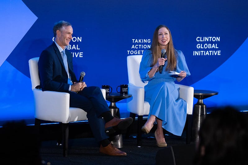 Chelsea Clinton and Tom Inglesby both sit in front of a blue background on white chairs.