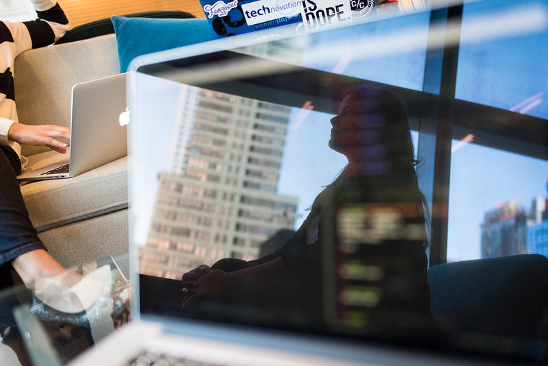 photo of a laptop computer screen showing a reflection of a person with long hair in profile against a city skyscraper background; another person sits on a couch behind the computer