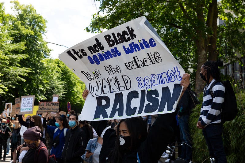 A person dressed in a black hoodie with a black face mask holds a hand-lettered sign above their head that reads, “It’s not Black against white. It is the whole WORLD against RACISM.” There is a crowd of people in the background, also holding signs above their heads.