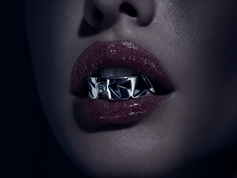Close-up beauty shot of glossy lips wearing a futuristic chrome grill accessory with sculpted metallic lettering in a dark cinematic aesthetic