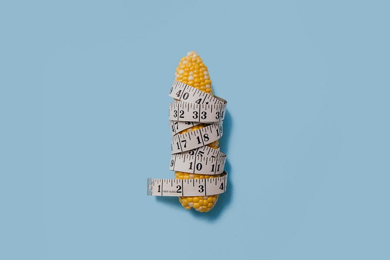 An image of a single corn on the cob wrapped in a measuring tape, in front of a baby-blue background.