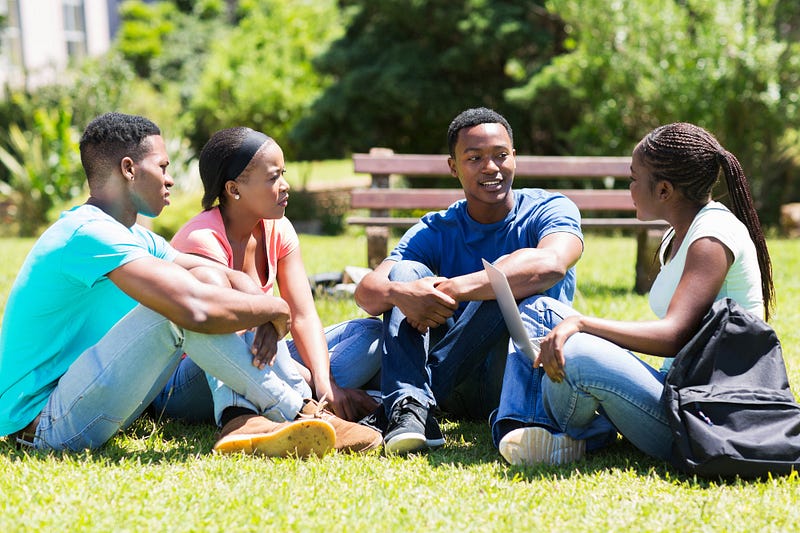 Image of a group of 4 students sitting with their legs crossed, chatting with each other on the grass. From left to right: young, masculine-presenting Black person with a cyan colored shirt, jeans, and tan shoes; young, feminine-presenting Black person with a black headband and coral T-shirt; young, masculine-presenting Black person with a dark blue shirt, jeans, and black shoes; young, feminine-presenting Black person with teal T-shirt, jeans, and white shoes, holding a laptop on her lap.