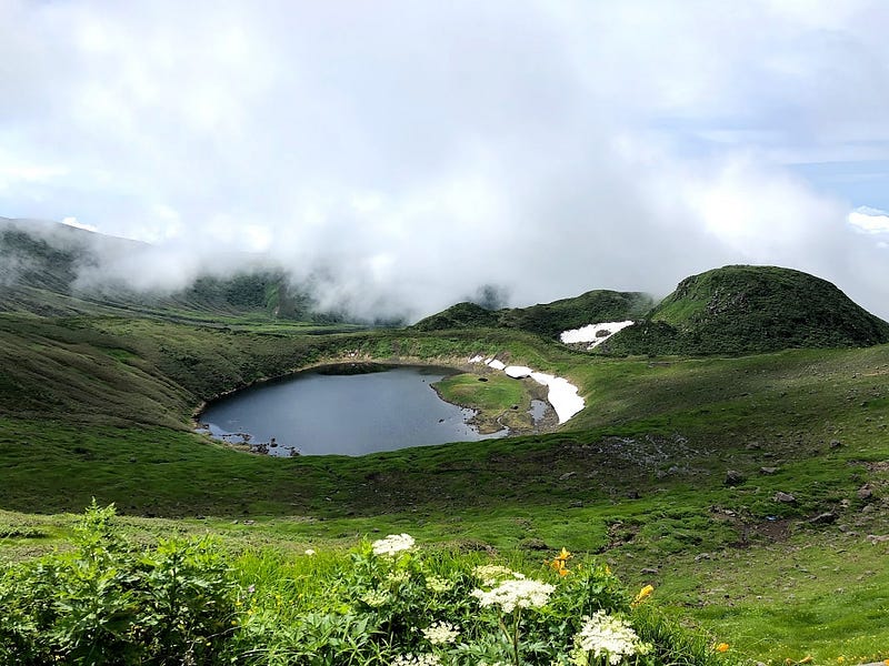 Chokai-san (Mt. Chokai)’s crater lake surrounded 360 degrees by green meadows and yellow dawn lilies with a giant white cloud above.