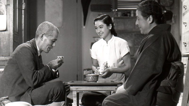 A smiling elderly couple and young woman sit cross-legged on the floor at a table. The man holds his cup up towards him.