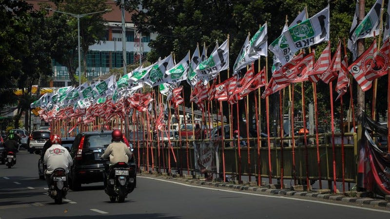 A wide angle photo of an Indonesian two way road with a median that has hundreds of PKB and Partai Kebangkitan flags. Two motorcycles and three cars can be seen moving away from the photographer in the lower left side of the image.