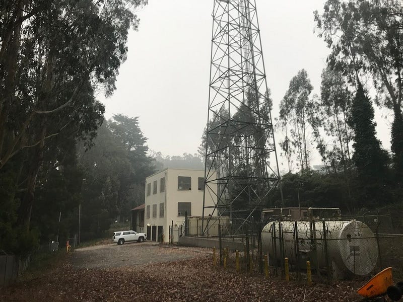 A small building next to a metal tower and a large tank, surrounded by trees and fog.