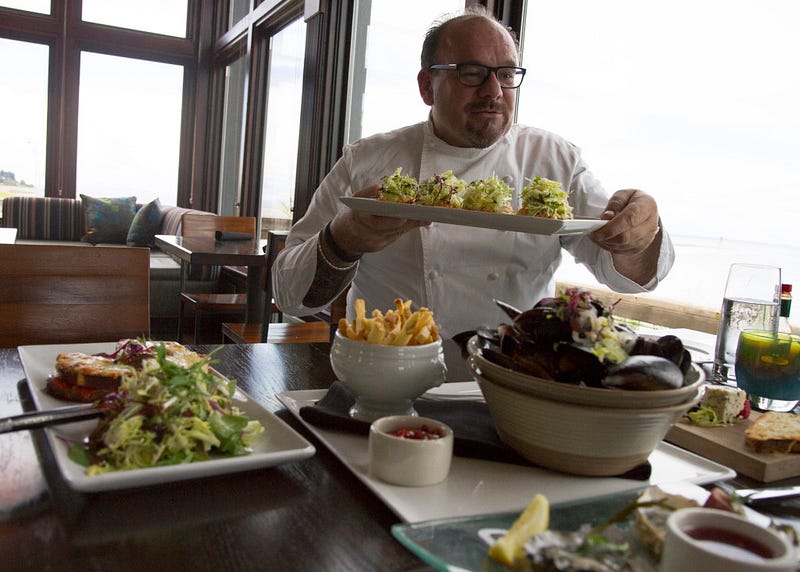 Chef Eric A. Truglas passes a plate of fresh house-made artisan bread topped with Dungeness crab, arugula and tomato vinaigrette in Packers Oyster Bar. Other dishes include steamed mussels from Whidbey Island’s Penn Cove Shellfish and a PNW take on the traditional grilled ham and cheese sandwich featuring house smoked salmon.