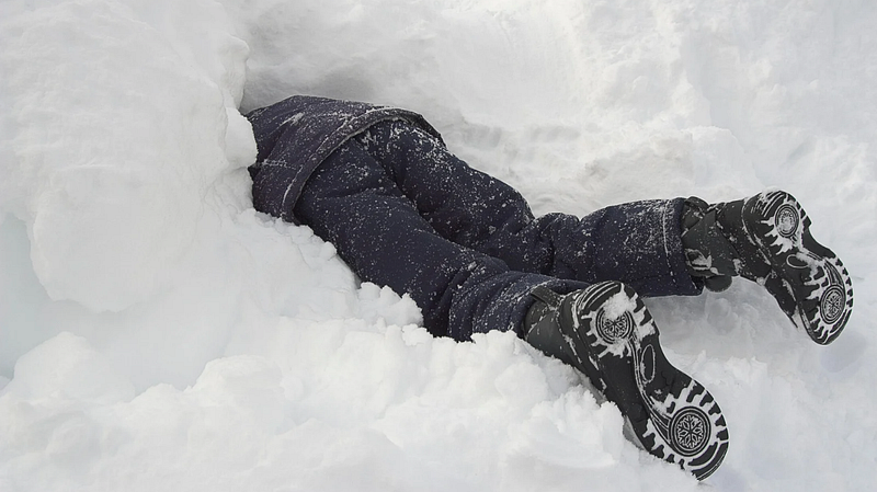 Person buried in snow — A person wearing a dark winter outfit is partially buried in snow, with only their legs and boots visible, lying face down.