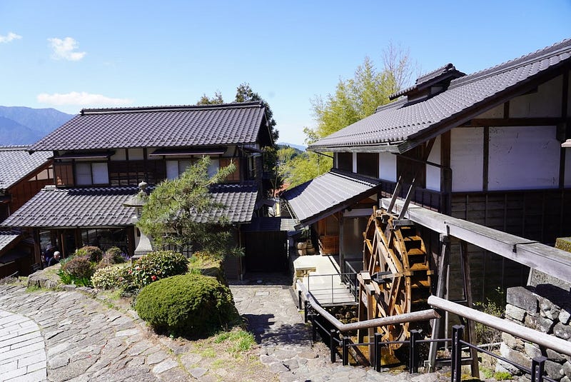 Waterwheel and buildings beside a sloping stone walkway overlooking mountains in the distance.