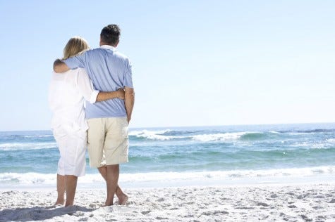 Senior Couple On Holiday Walking Along Sandy Beach Looking Out T