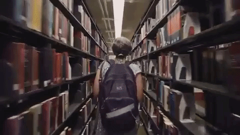 Gif de uma mulher branca de cabelos grisalhos, com uma mochila nas costas, andando em um corredor de uma biblioteca e depois conversando com um homem preto, de óculos, vestindo uma camiseta na cor branca.