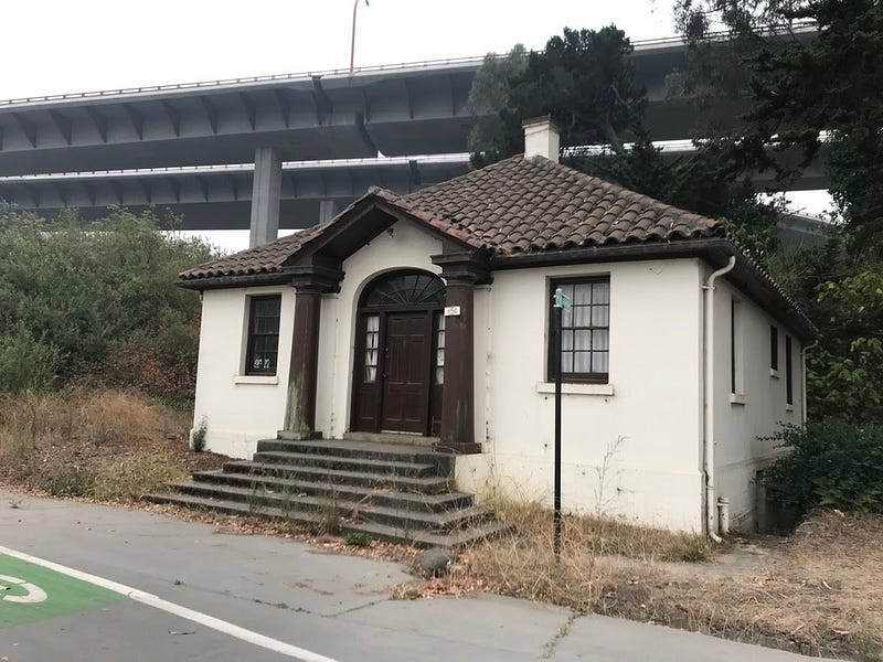 A small, single-story house-like building with freeway bridges behind it.