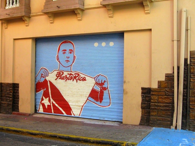A photo of a mural on the side of a building. The mural’s background is light blue, and there is an outlined picture of a man holding a shirt or fabric that says Puerto Rico and has part of the Puerto Rican flag.