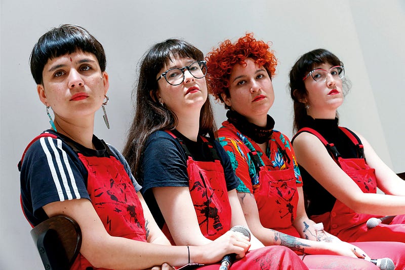 Image description: four members of feminist performance art group, Las Tesis, sitting in a row wear red and black with red overalls. their bodies face the observer’s right, but their heads are camera-facing. two individuals hold a microphone on their lap